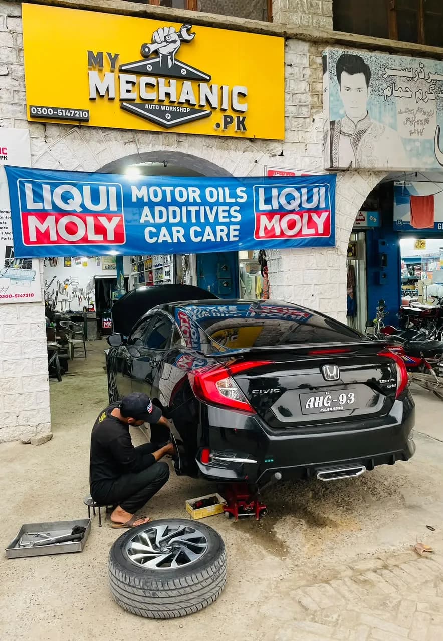 Technician working on a black Honda Civic raised on a jack with one wheel removed, outside the My Mechanic shop