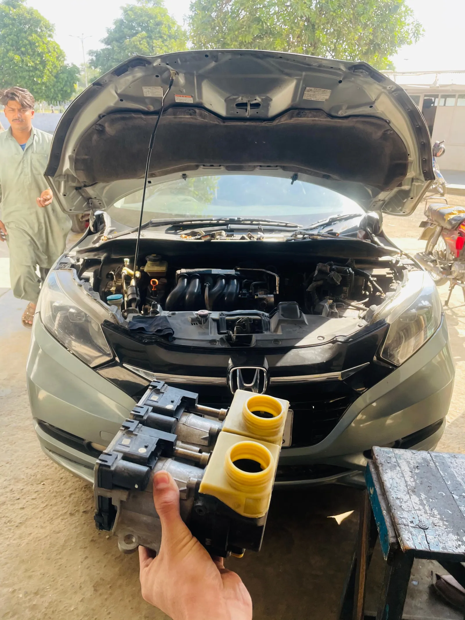 Mechanic holding a dual-clutch transmission control unit in front of a Honda with its bonnet open