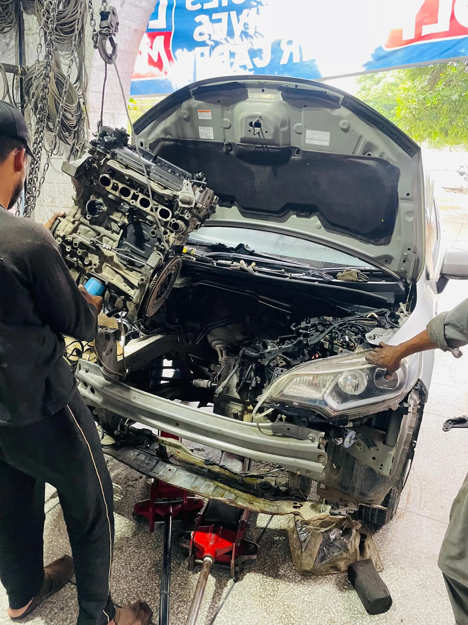 Mechanics lifting the engine out of a car with its front end stripped, during an engine overhaul