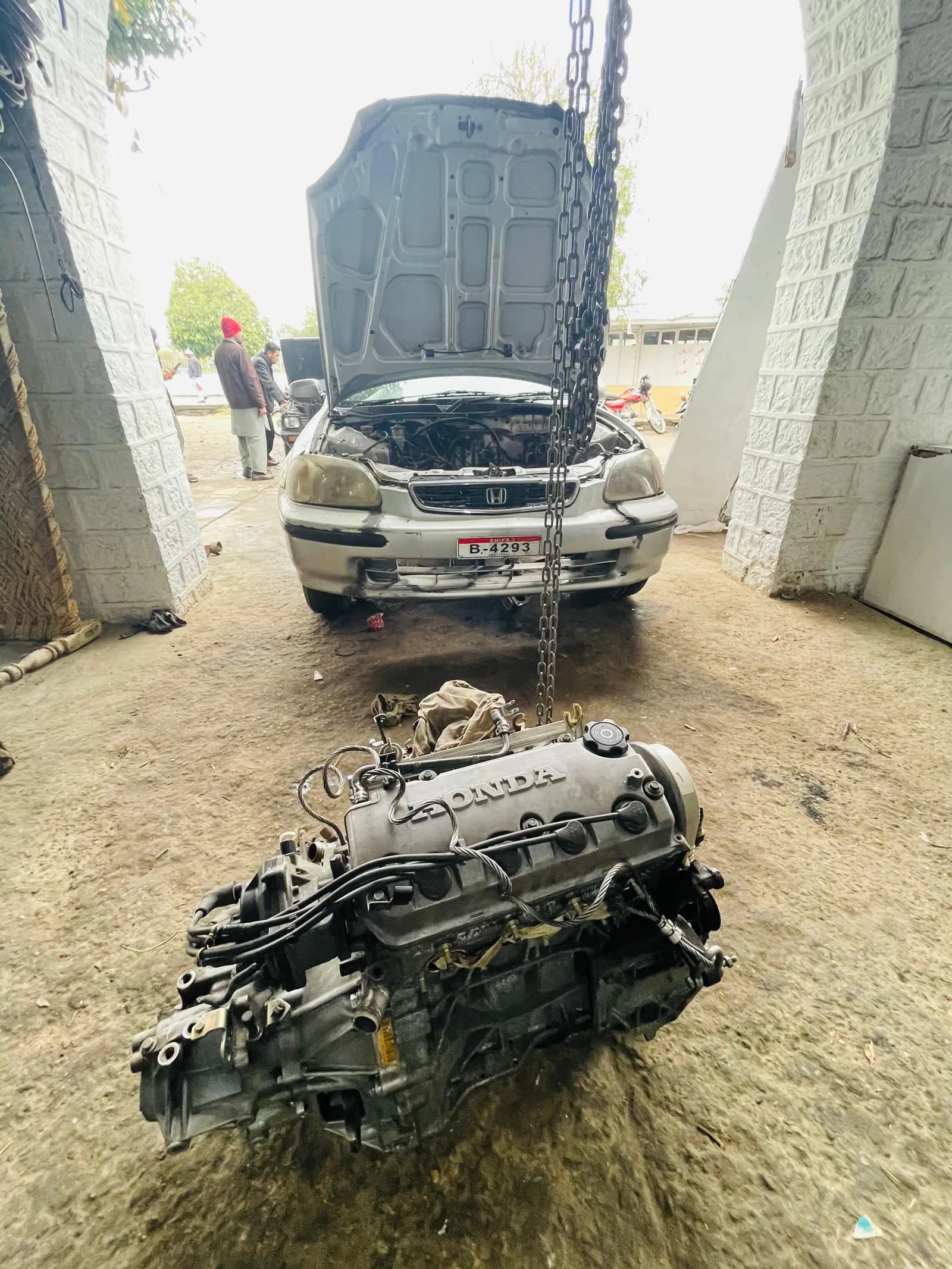 Replacement engine on the workshop floor in front of the car it is being fitted to, with a chain hoist above