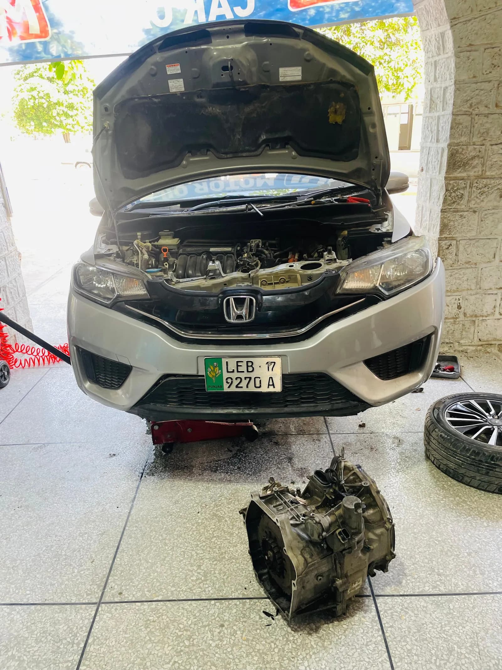 Honda Fit hybrid raised for repair with its gearbox removed and placed on the floor in front of the car