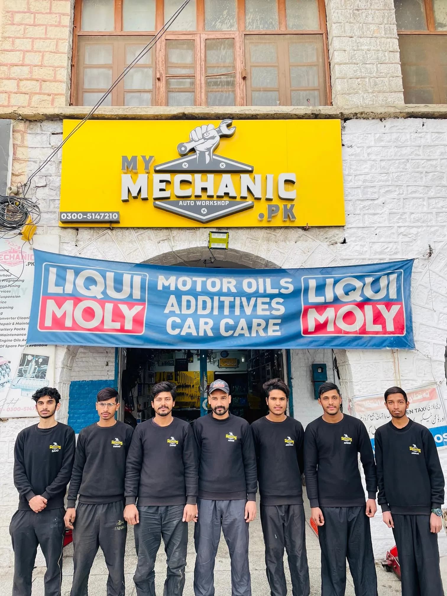 The My Mechanic team in uniform standing in a line in front of the workshop's yellow sign and Liqui Moly banner